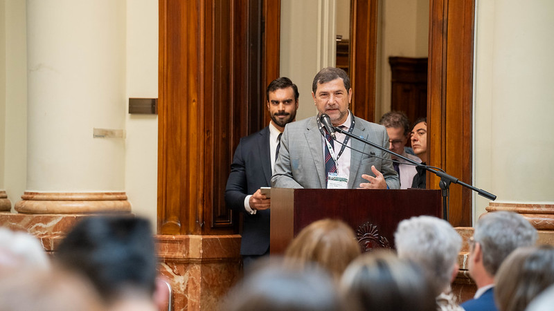 Photo of a man in a grey suit speaking at a lectern, with the backs of people's heads in the foreground