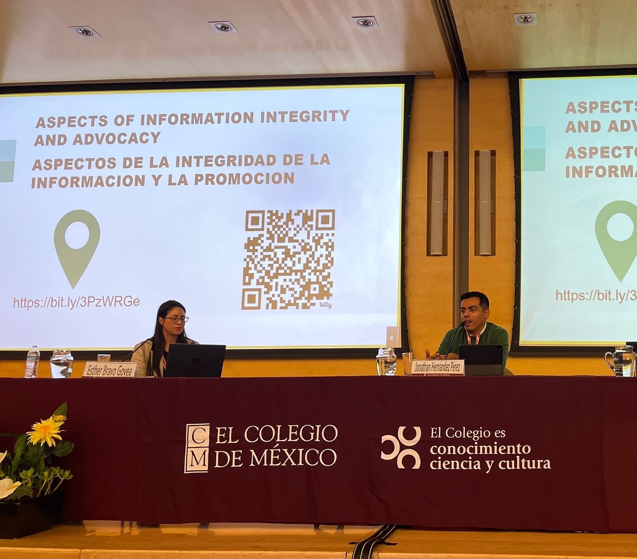 Two people on a podium at an event. The table in front of them includes the logo of the Colegio de Mexico, and behind there is a slideshow wiht a QR code and question about what a healthy information environment looks like