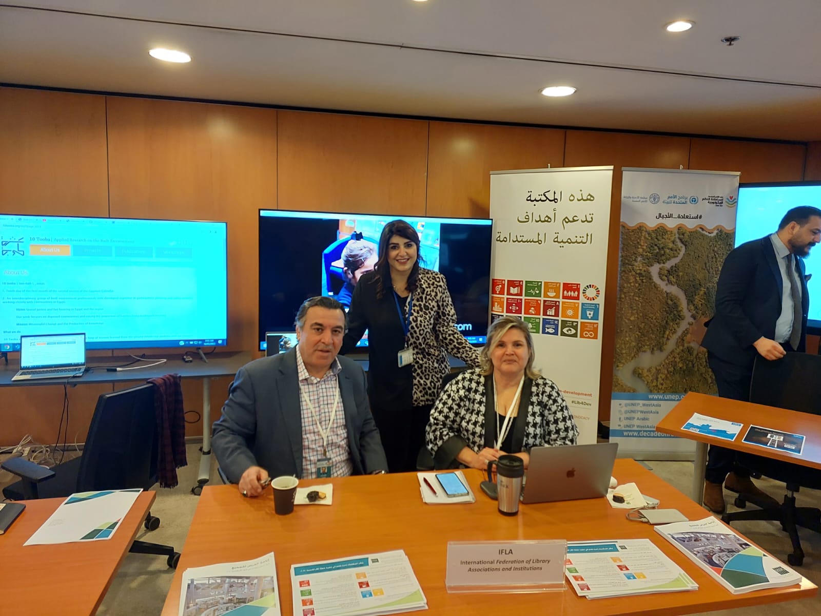 AFSD ESCWA with MAYA from the ESCWA Library Photo of three people sitting at a table in a conference centre, with banners and screens behind them, and materials on the table in front of them