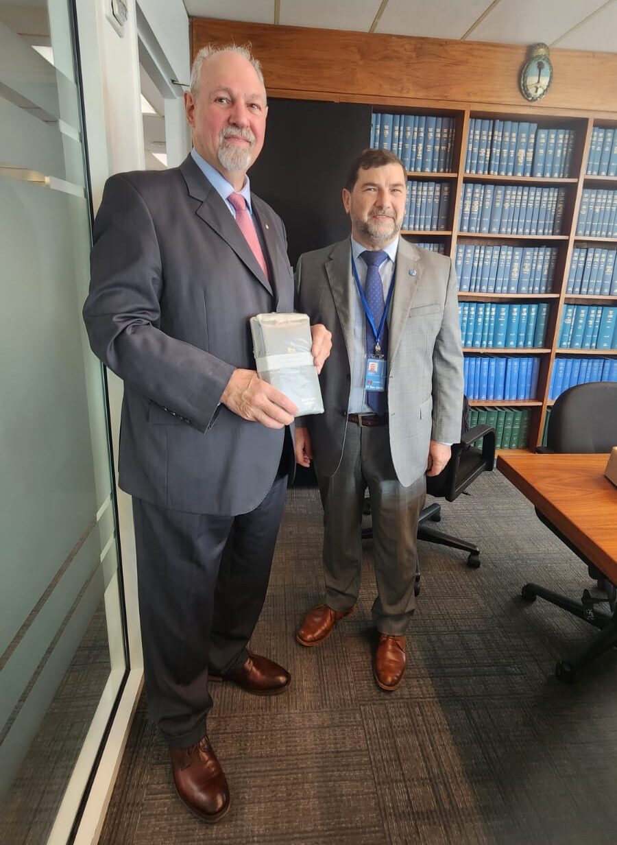 Two men in suits in a meeting room, with a bookshelf in the background. The man on the left is holding a book in a wrapper that has been presented to him