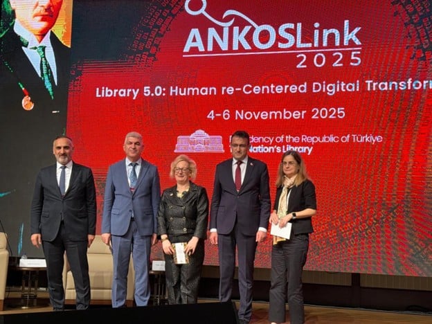 A group of five people stands in front of a presentation about Library 5.0: Human re-centered digital transition. The man on the right is in a dark suit, white shirt and grey tie. He has a beard and has a receding hairline. The next man has a light grey suit, white suit and grey tie. He has grey hair. The woman in the middle has Three people stand for a picture in front of a background with ANKOSLink and English and Turkish text. The man is on the left in a brown leather jacket, a black shirt and trousers and is balding with glasses. The woman in the middle is in a black suit, is blond and has glasses. The next man is in a dark suit, white shirt and red tie, and has dark hair. The woman on the right is in a black top and trousers, and a white shirt. She has glasses and brown hair.