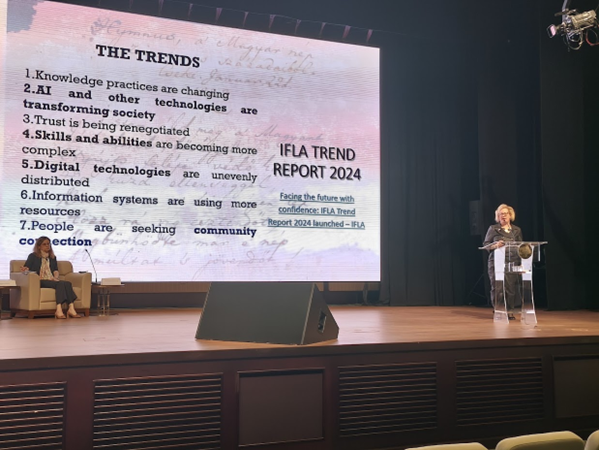 A blond woman stand at a lectern giving a presentation. On the big screen behind her the IFLA Trends are listed. A woman looks on from an armchair.