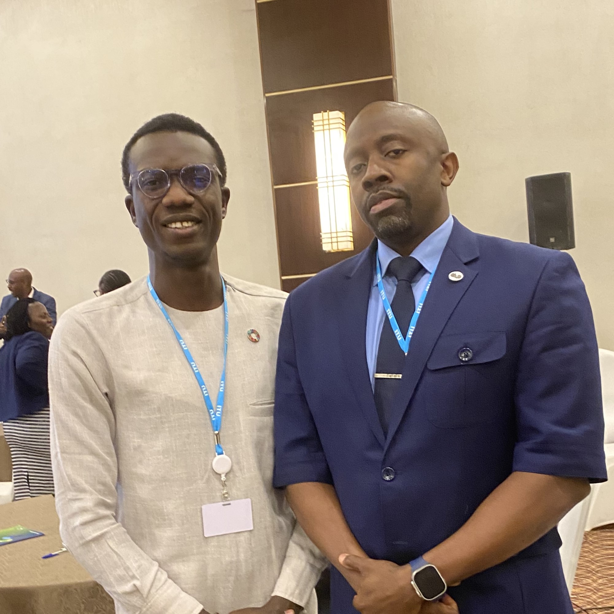 Damilare Oyedele with Choolwe Andrew Nalubamba Director General of Zambia Information and Communications Authority ZICTA Two African men. On the left a younger one in a beige top with an SDGs badge. On the right, an older one in a blue suit and tie.