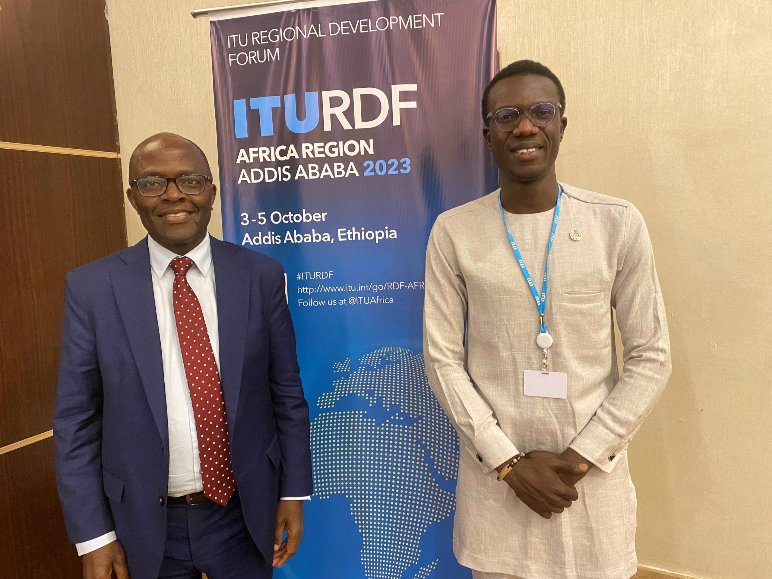 Damilare Oyedele with Dr. Cosmos Zavazava Director Telecommunication Development Bureau BDT International Telecommunication Union scaled Two African men. On the left, a smiling man with a dark blue suit, white shirt and red spotted tie. On the right, another smiling man in a beige top. Behind them, a banner with ITURDF and an image of Africa