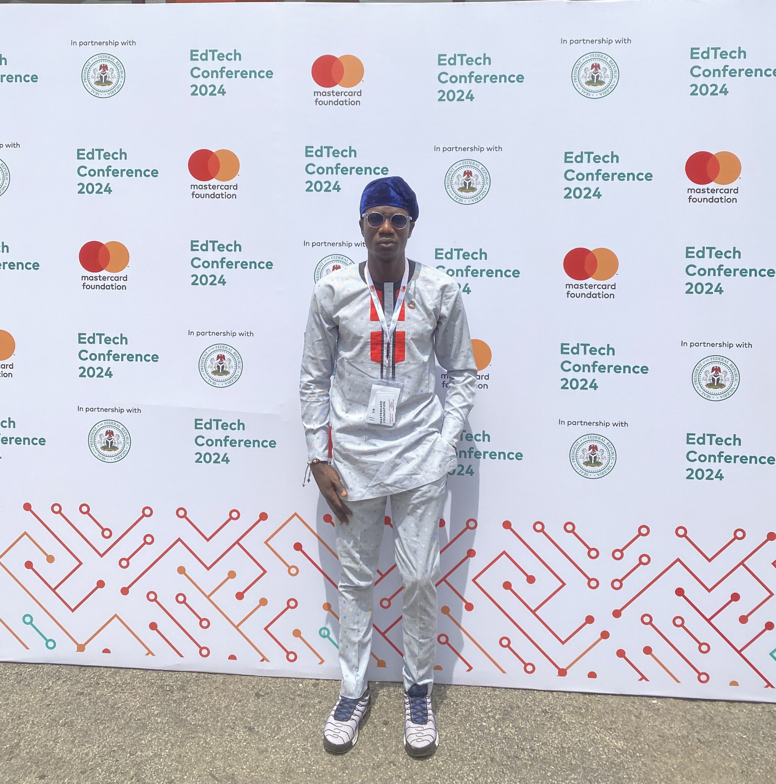 Man in a shiny white long-sleeved top and trousers in front of a branded background with Mastercard Foundation and EdTech Conference 2024 repeated across it
