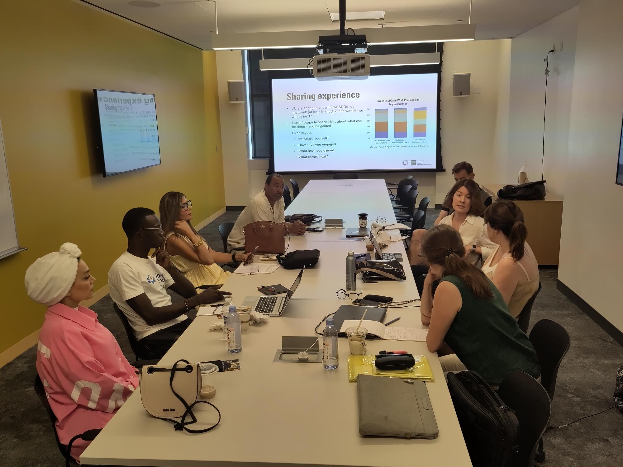 HLPF Workshop Group of people sitting around a table with a powerpoint in the background on a screen