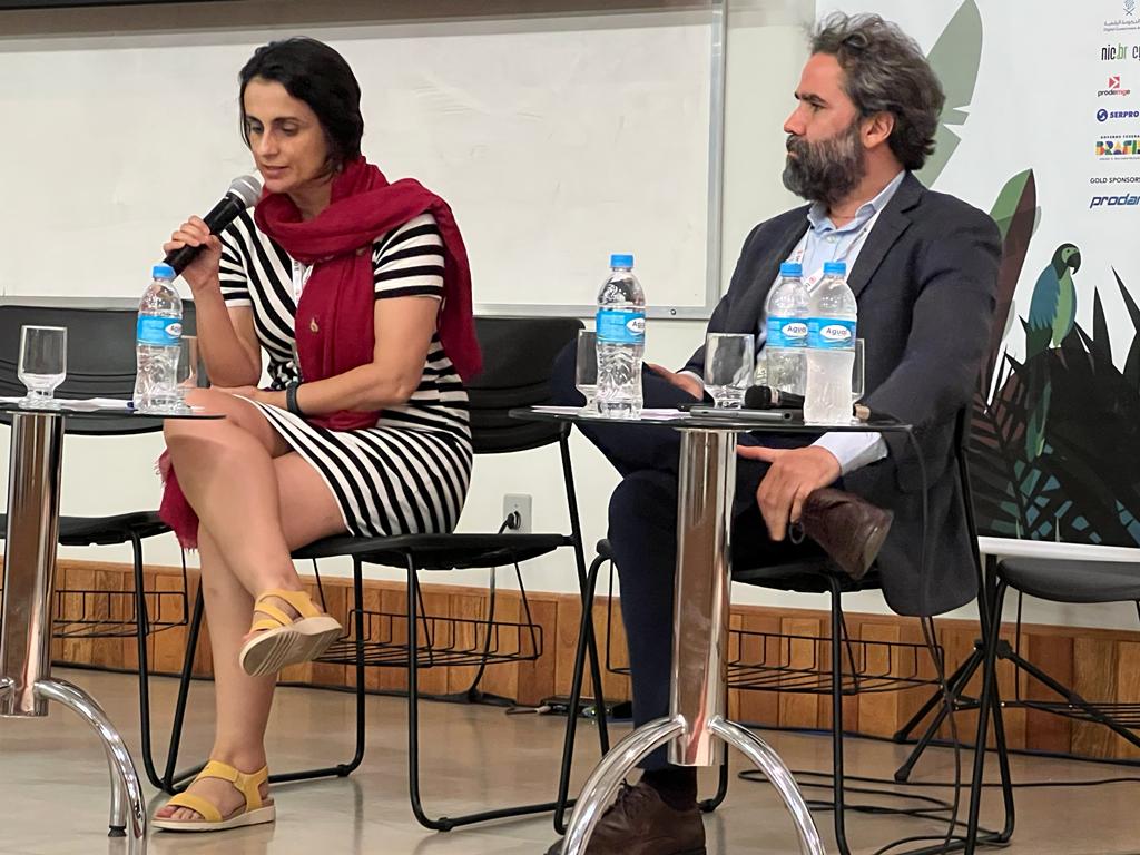 ICEGOV CleideFernandes 1 Photo of two people - a woman with a black and white striped dress and a red scarf (speaking into a microphone), and a man in a suit and white shift