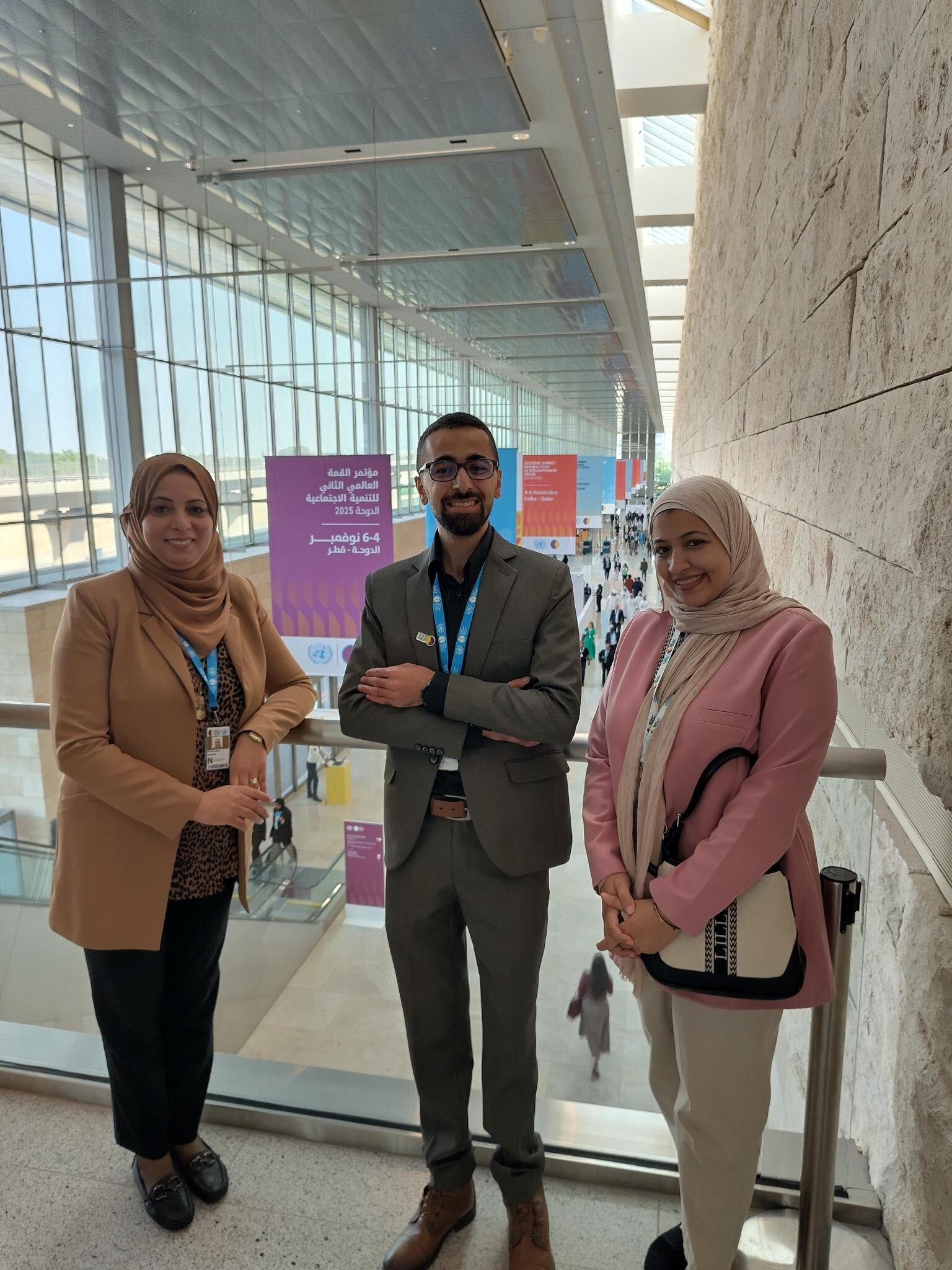 Group of three people standing on a balcony, looking down at an atrium in a conference centre. Banners for the Second World Summit on Social Development hand in the background, and there is a large plate glass set of windows looking to the outside. The woman on the left is in a beige jacket and headscarf. The man in the middle is in a grey-brown suit with a black shirt. He has a beard and is smiling. The woman on the right has a white headscarf and pink jacket, and is carrying a white and black handbag. They are all smiling. 