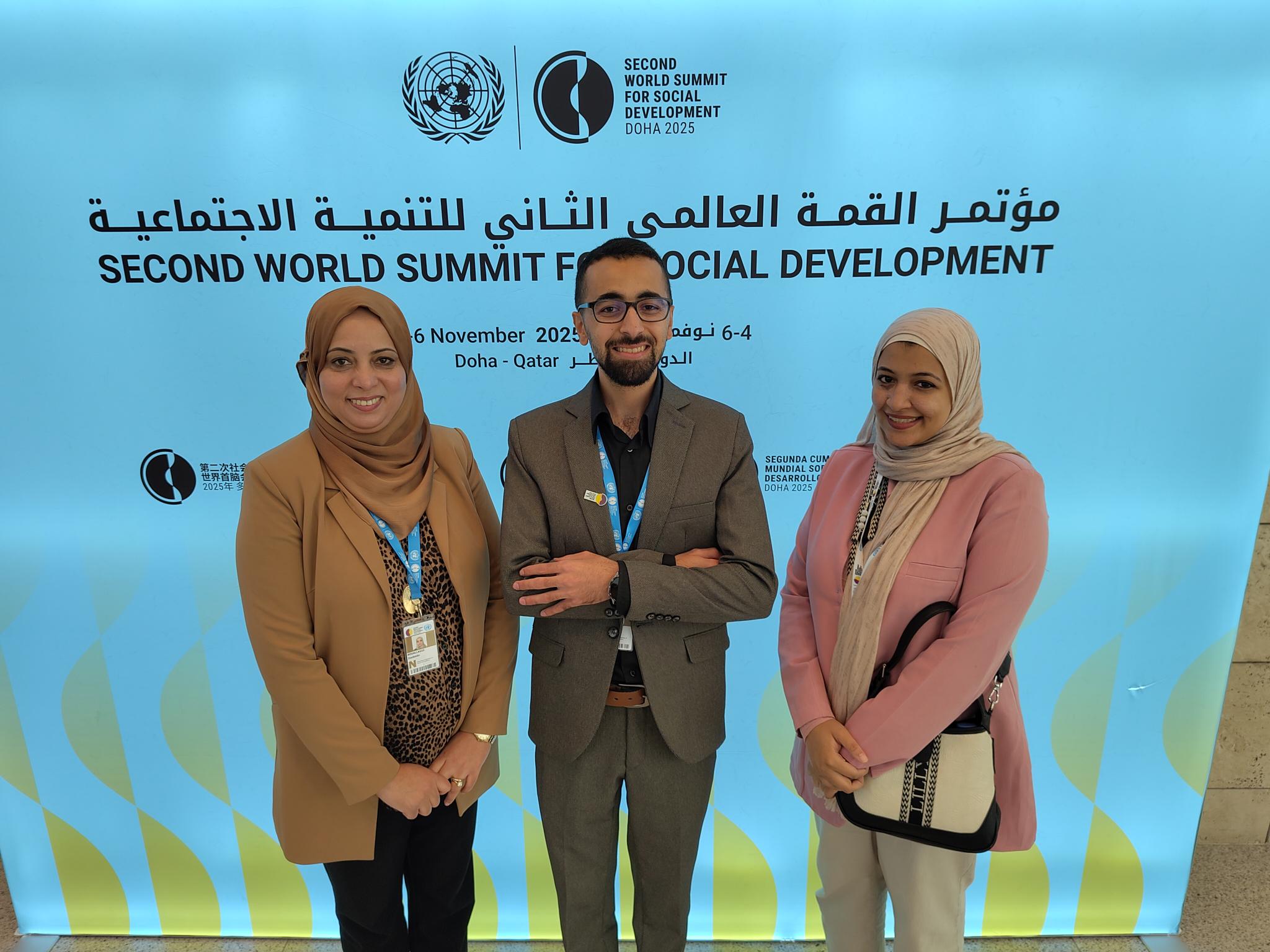 Group of three people standing in front of a back-lit blue background, with Second World Summit on Social Development written on it. The woman on the left is in a beige jacket and headscarf. The man in the middle is in a grey-brown suit with a black shirt. He has a beard and is smiling. The woman on the right has a white headscarf and pink jacket, and is carrying a white and black handbag. They are all smiling. 