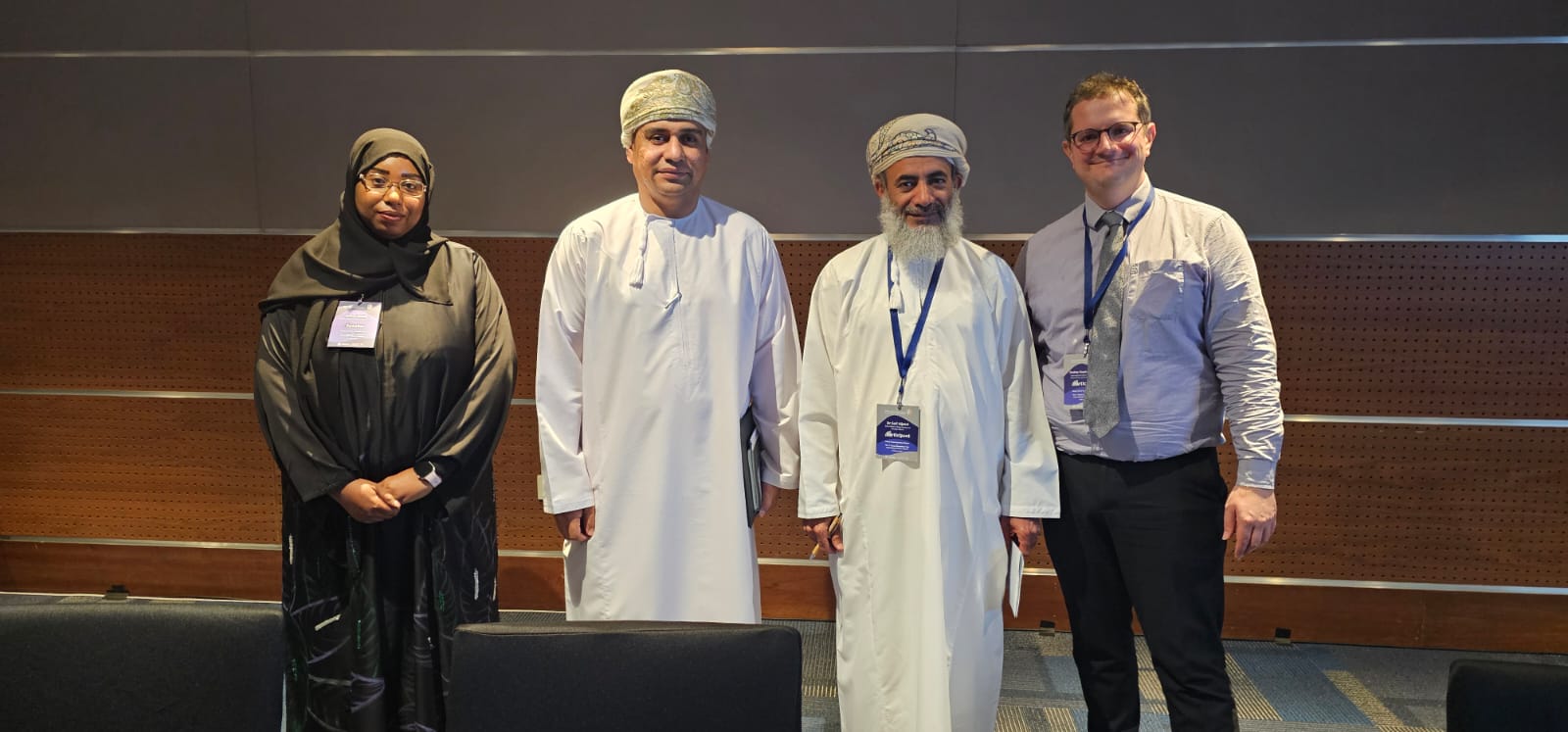 Four people standing in a line for a photo. A woman in black islamic clothing on the left, two men in Omani dress in the middle, and a white man in a shirt and tie on the right