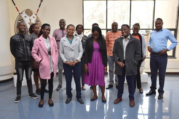 Group of Kenyan wikimedians, with a row of three women and one man in front, and another row of three women and five men behind. There is a window and a traditional shield and asegais in the background