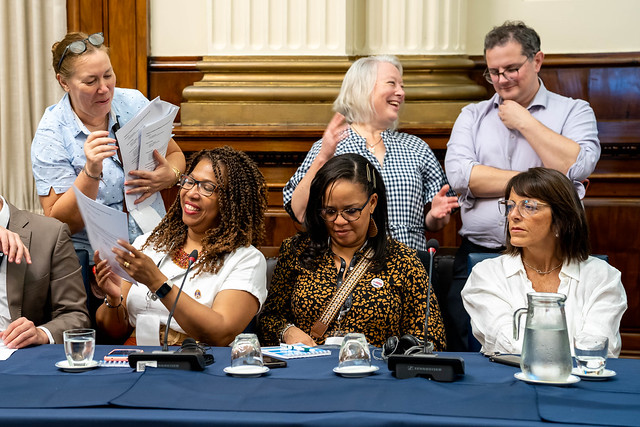 Three people sitting at a table, talking, looking at a phone and listening, three others behind chatting and working