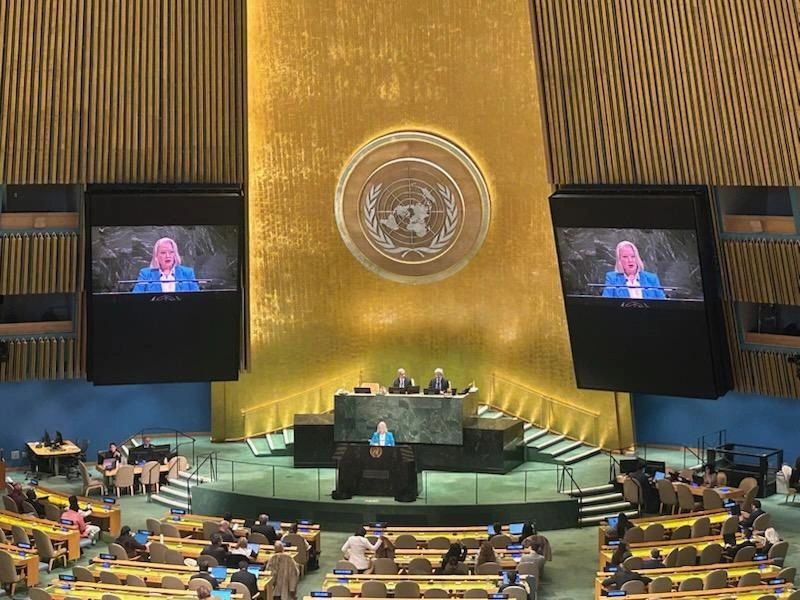 UN General Assembly Hall. A podium with a lectern at the front, with a golden wall behind and a UN logo. A woman with grey hair and a turquoise jacket is speaking, projected onto two screens on the wall