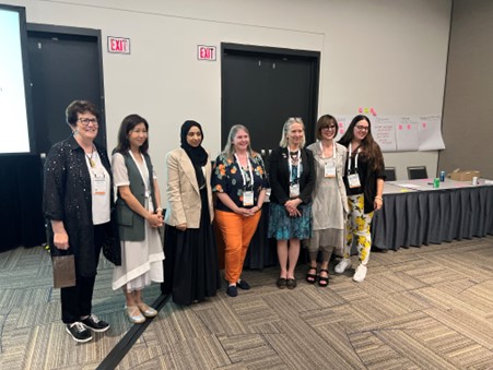 Group of smiling speakers at a conference event, standing in front of a table with a wall and door in the background.