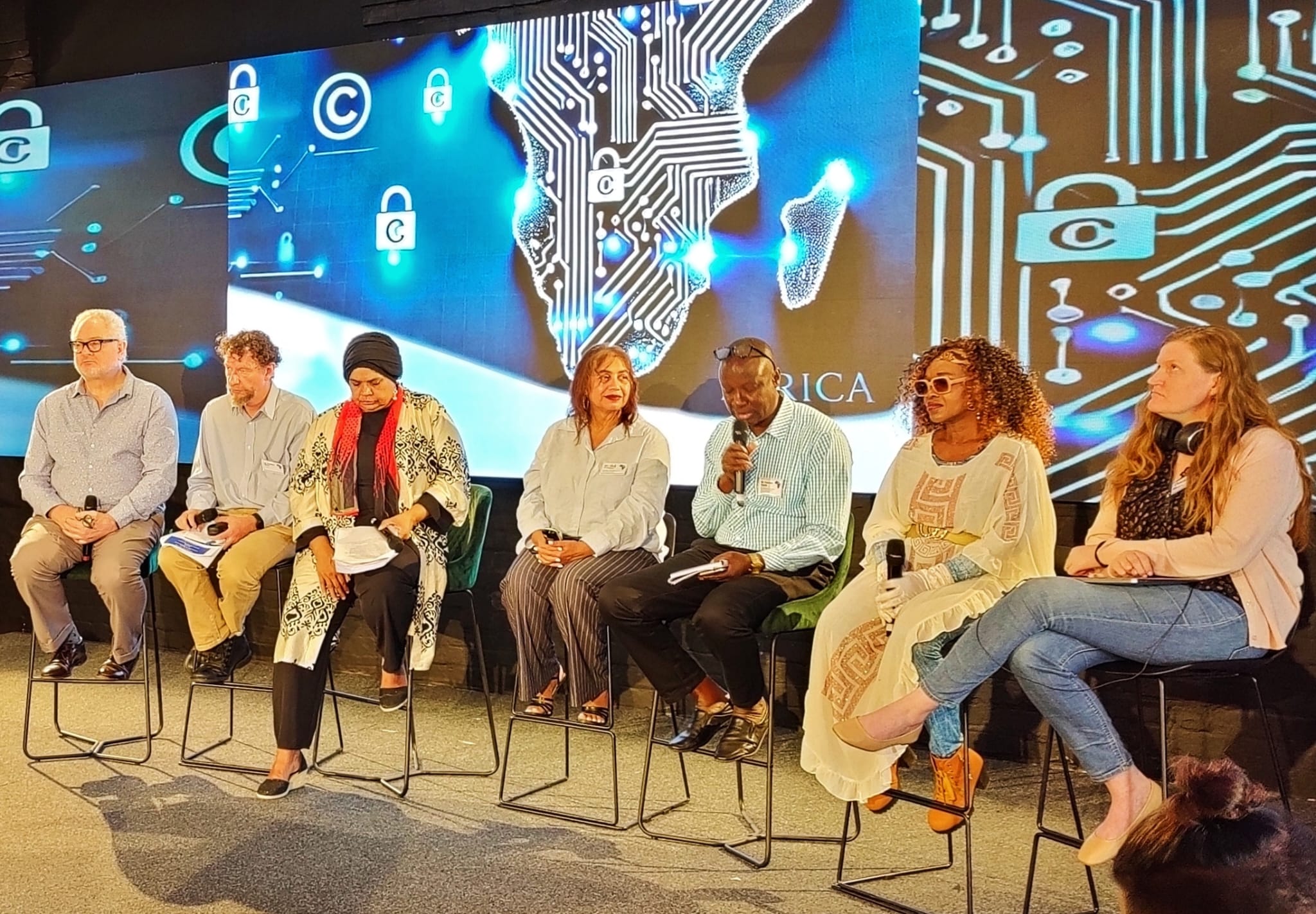 Group of people on bar chairs on a stage, talking to an audience, with an image of Africa made up of a circuit board behind them