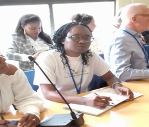 Woman with a white t-shirt, blue lanyard, mid-length curly black hair and glasses, sitting at a desk with a microphone and other people around her