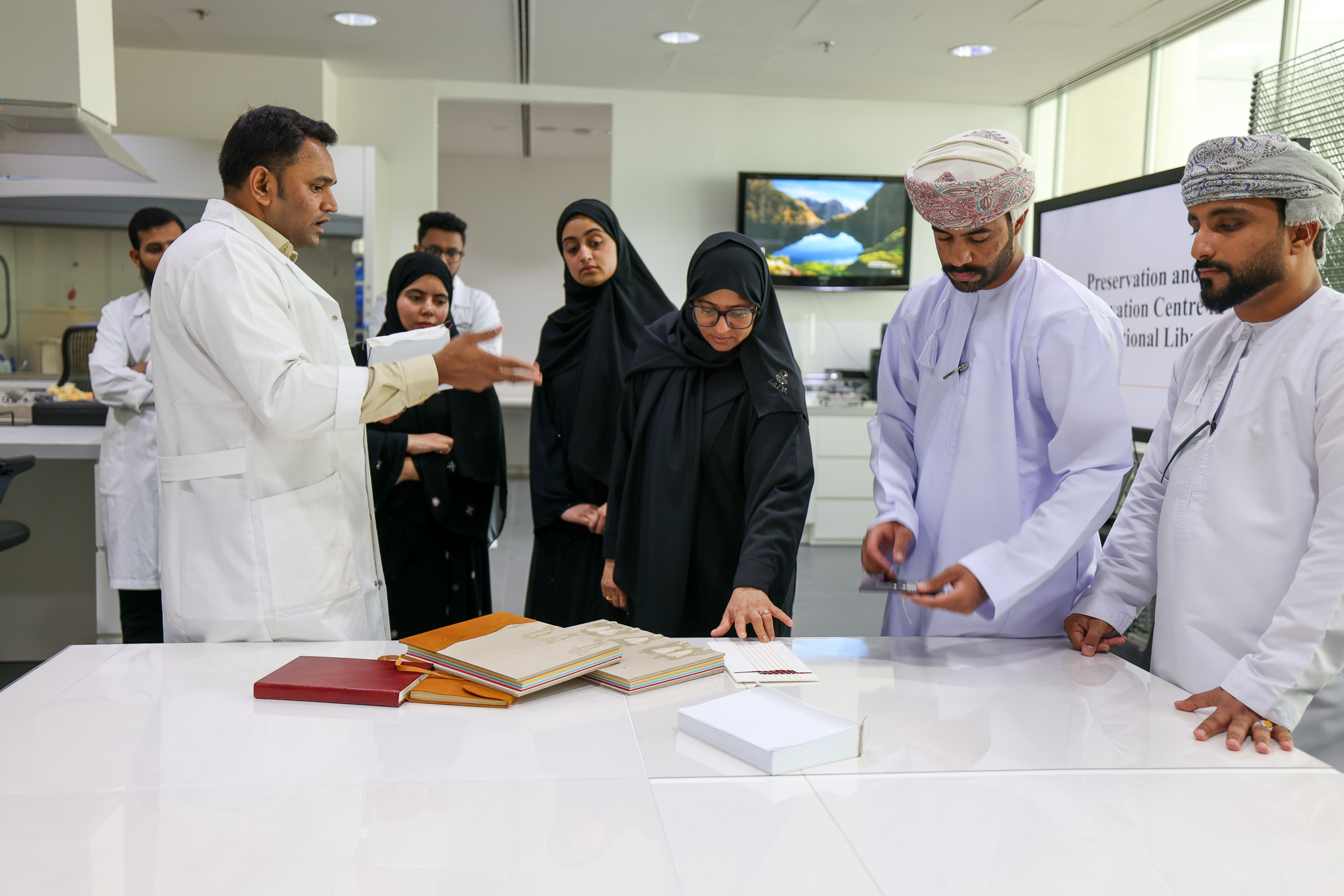 Group of people in a preservation lab. A man in a lab coat is explaining something to a group of women and men (women in black with headscarves, men in white with turbans). On the table, there are some books.