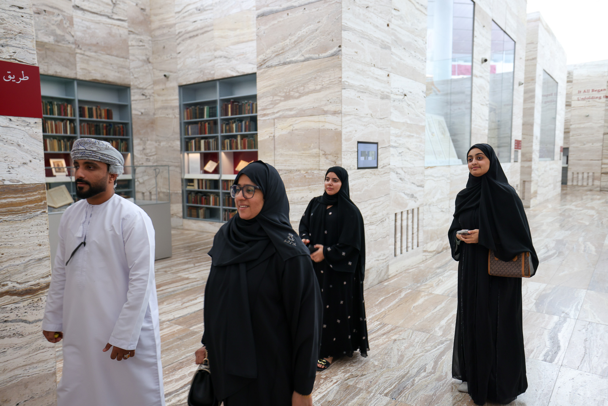 Group of people in a building with bookshelves and marble walls. One man in white with a turban, three women in black, with headscarves