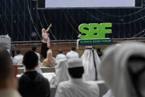 A woman in a hijab presenting on stage at the Science Book Forum with an audience seated facing her.