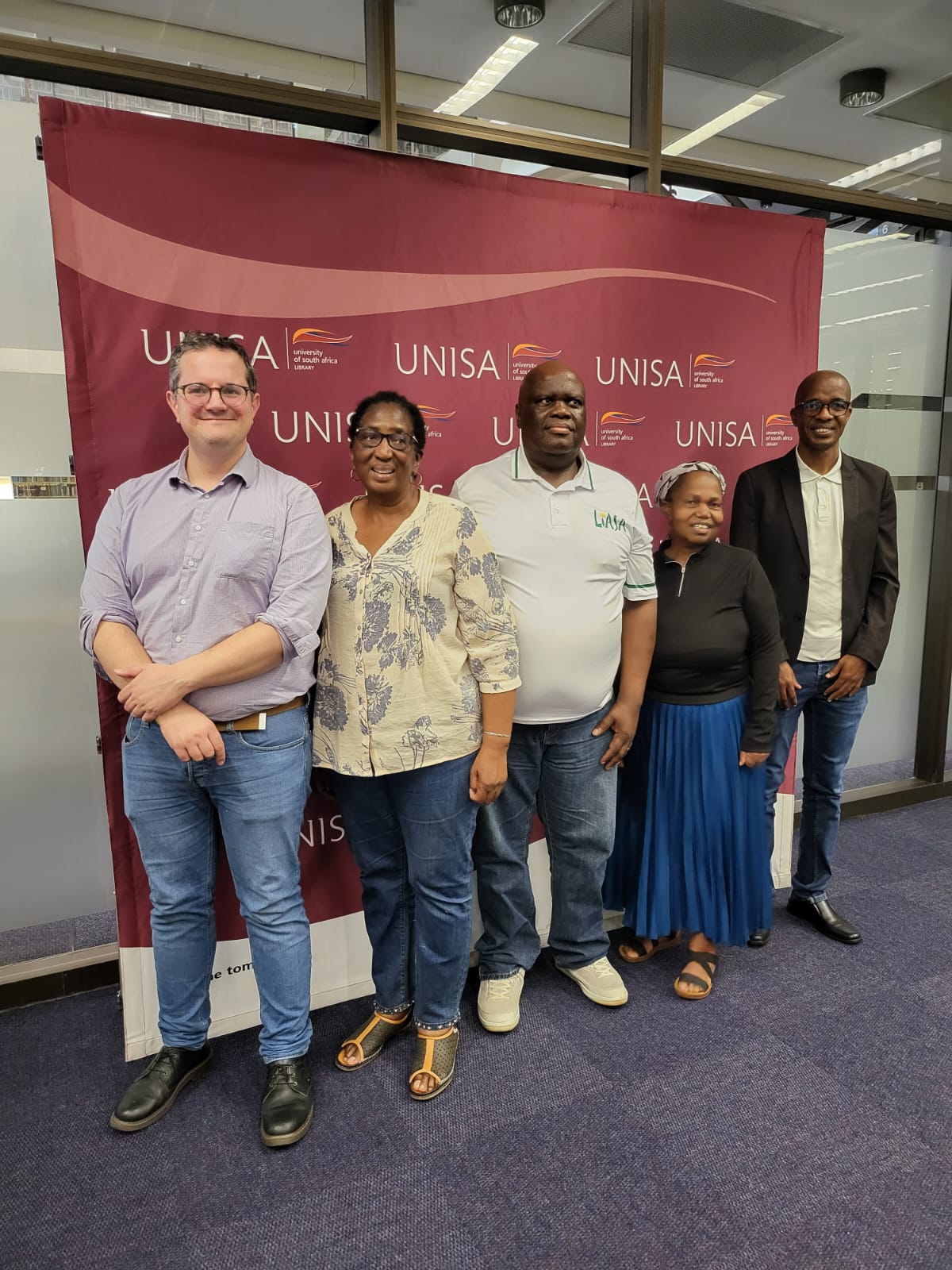 Group of four people (one white, left, and four black African to the right, with a UNISA banner behind)