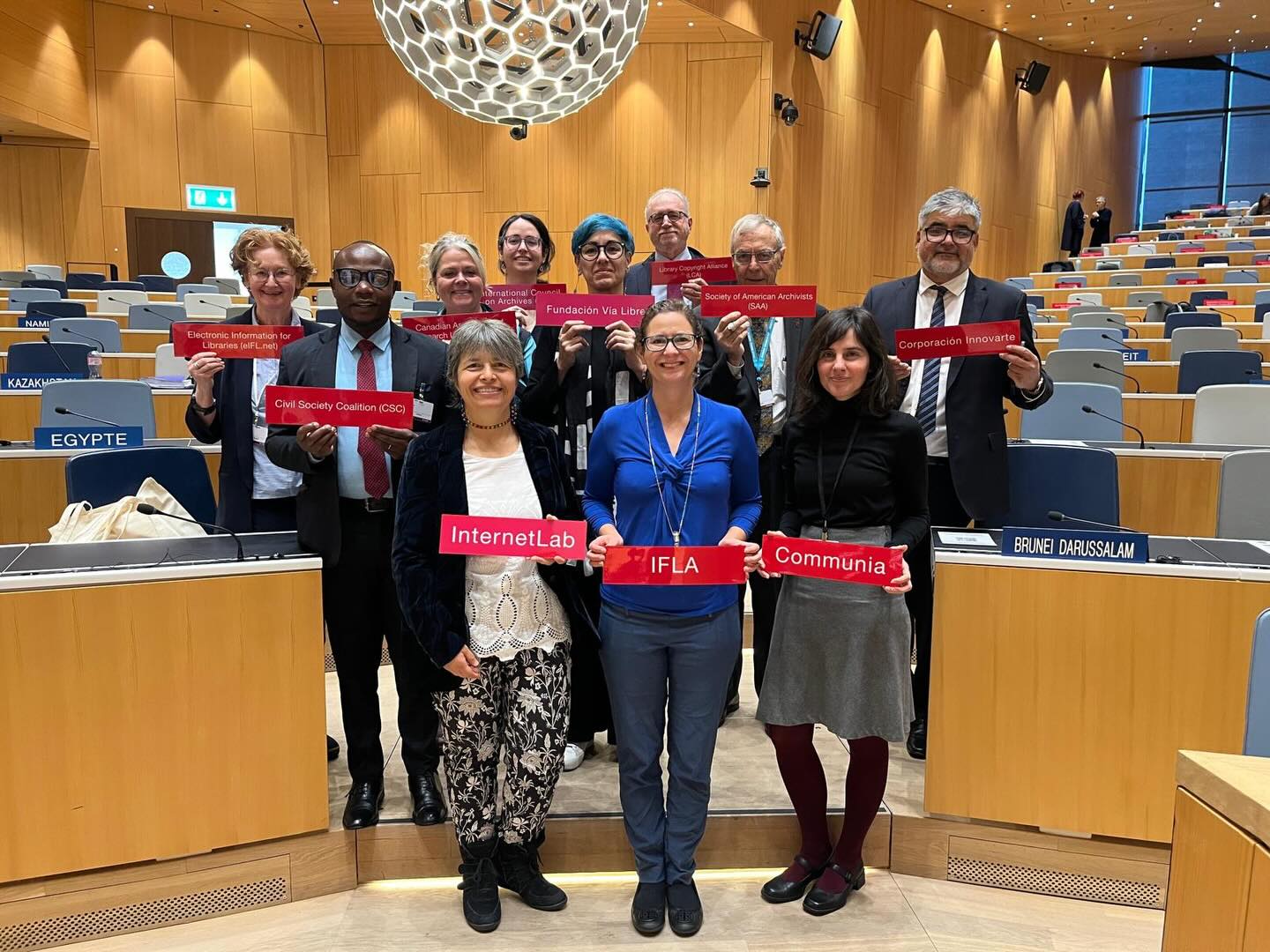 Group of people in a modern wood-lined room, holding up the names of their organisations on red plates