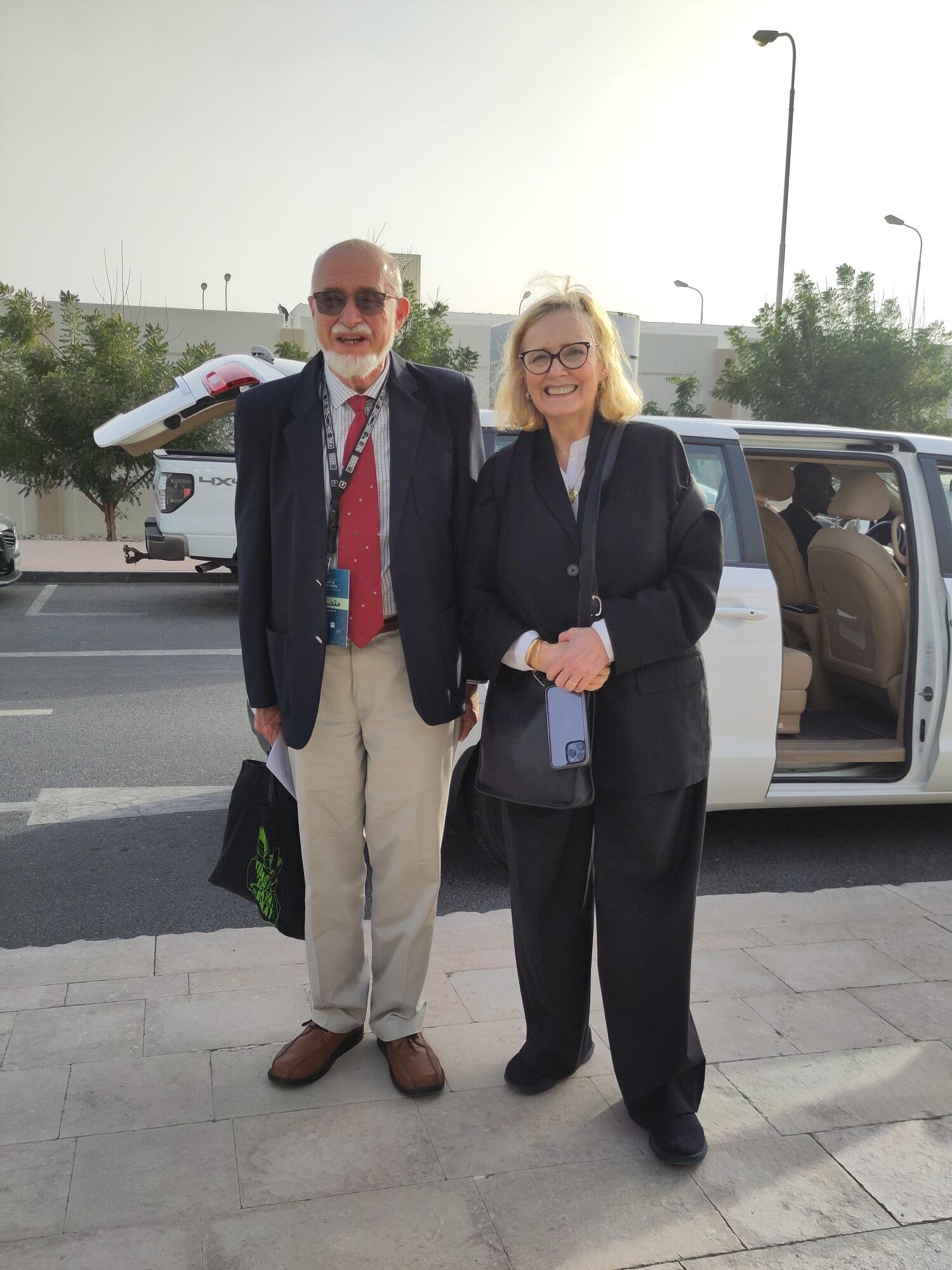 A white man with a white beard and bald head in a red tie, blazer and khaki trousers, and a white woman with blond hair, a loose black suite and a blue scarf. There is a car in the background