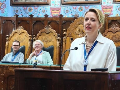 In the foreground a woman with blonde hair and a white shirt speaks into a microphone. Behind, two smiling people (a man with short dark hard and a blue shirt and a woman with short blonde hair, a light green jacket and a white top) sit on elaborate wooden chairs. The lower wall behind is of dark wood, and above is a blue painted wall with folk art patterns.