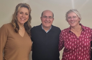 Picture of three smiling people, waist-up, against a white background. On the left is a taller woman with medium-long blond hair, and a beige top. In the middle is a balding man with glasses, a light blue shirt and dark blue pullover. On the right is a woman with shorter blonde hair, glasses, and a red shirt with white patterns.