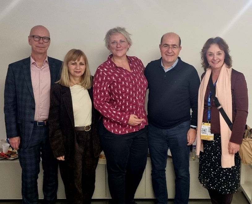 Photo of four smiling people against a white background. The man on the left is bald with glasses, a dark blue checked jacket and trousers and a light pink shirt. The next is a shorter woman with blonde hair and a dark suit and white top. The next is a taller women with short blond hair, a red shirt with white patterns and black trousers. The next is a balding man with glasses in a blue shirt, and dark blue pullover and jeans. The final is a woman with shoulder-length brown hair, a brown shirt, a pink scarf, and a black and white skirt.
