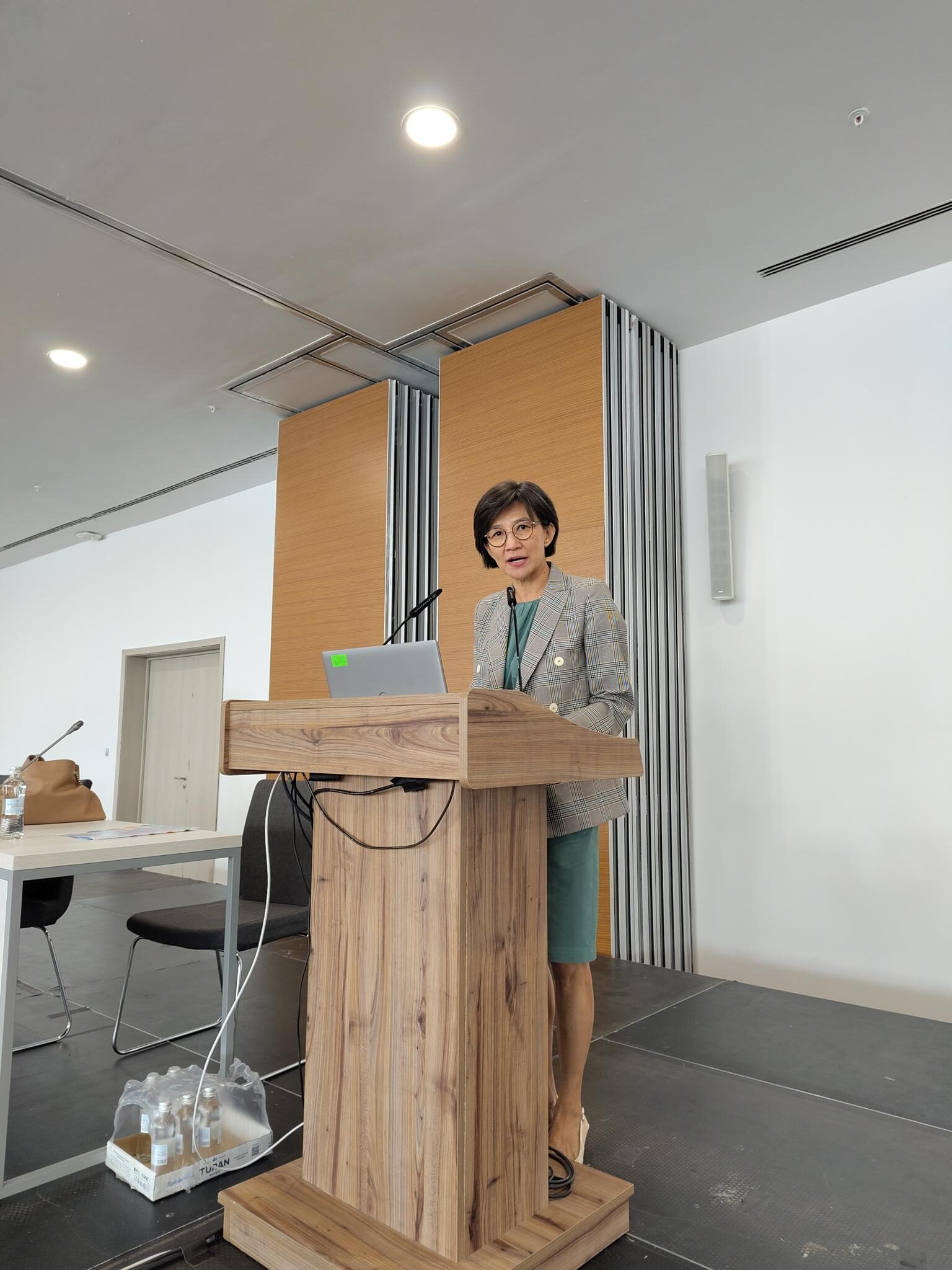 Photo of a woman with shortish dark hair, glasses and a grey suit speaking at a lectern