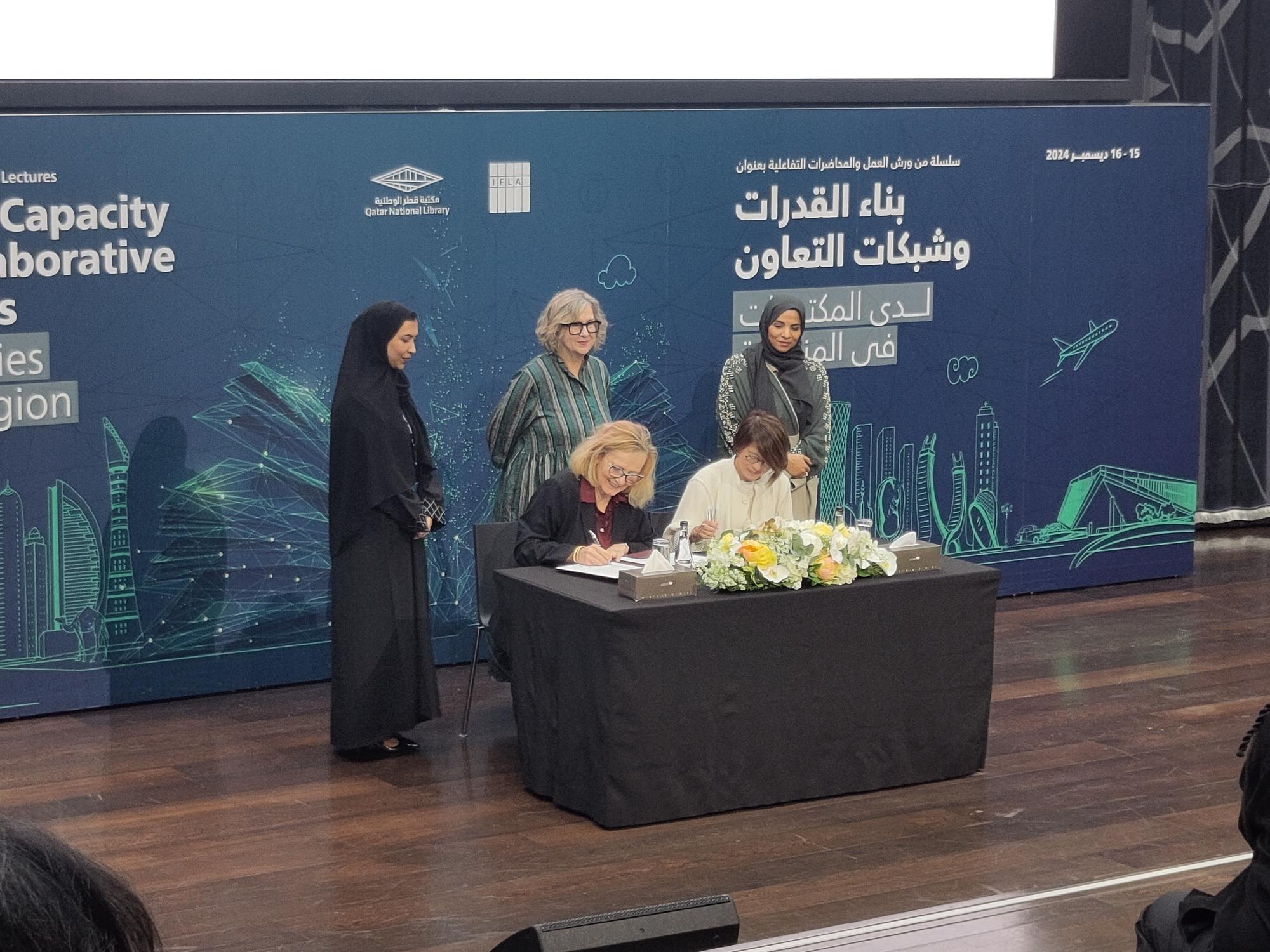 Two women sit at a table with flowers and a black tablecloth signing a document. Behind them, a woman in islamic dress, a blond woman in a green dress, and another woman in islamic address.