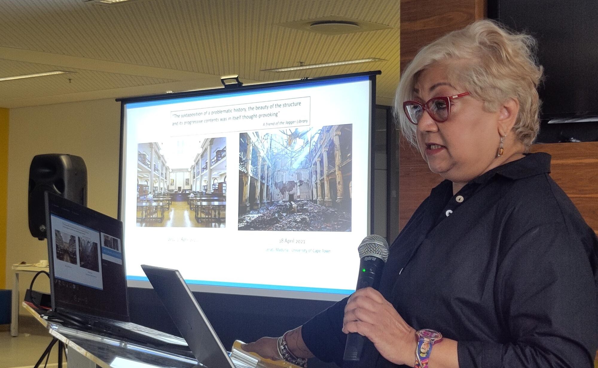 Woman with glasses and light blonde hair speaking into a microphone, with, on a projecting screen behind her, pictures of a burnt-out librari