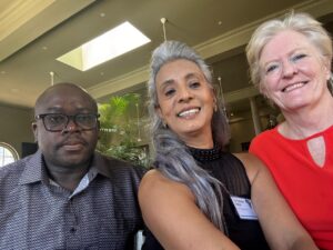 Photo of three people - one black African man with a grey shirt and glasses, one Indian South African woman with long grey hair and a balck top, and one white woman with short blonde hair with an orange top
