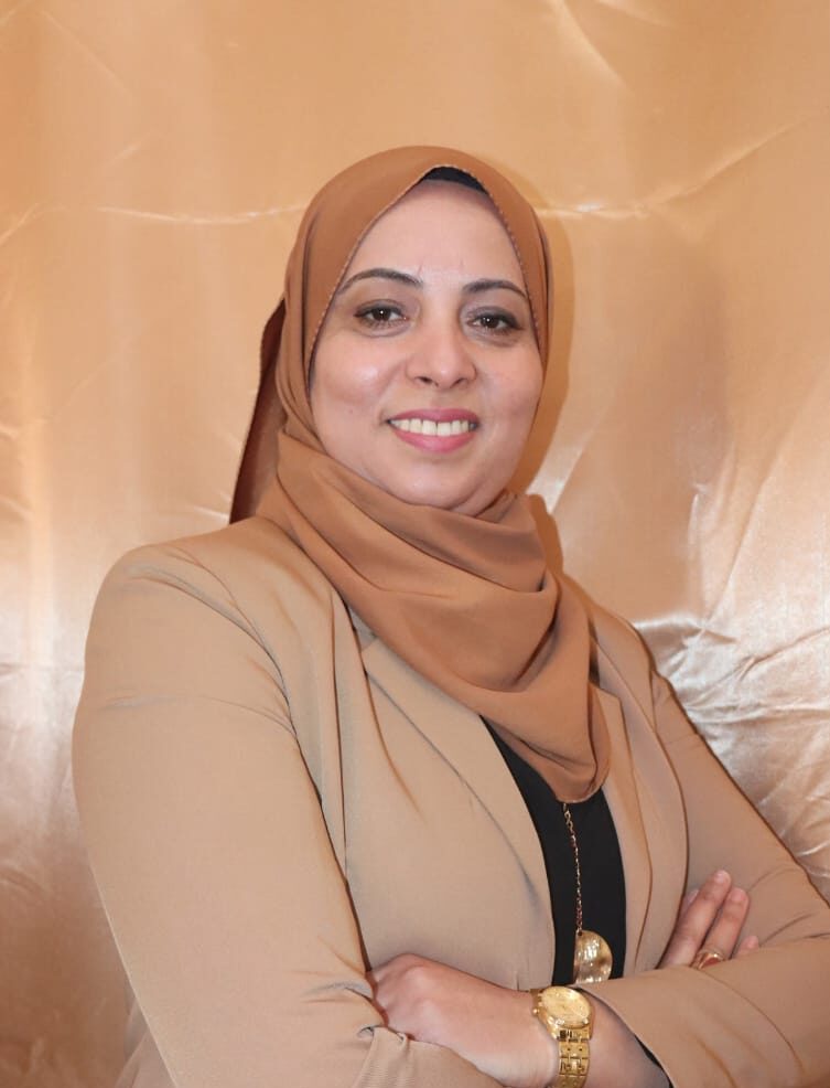 Photo of a smiling Tunisian woman in a light brown headscarf, and a lighter brown jacket, with a black top underneath. She is standing against a wooden wall background.