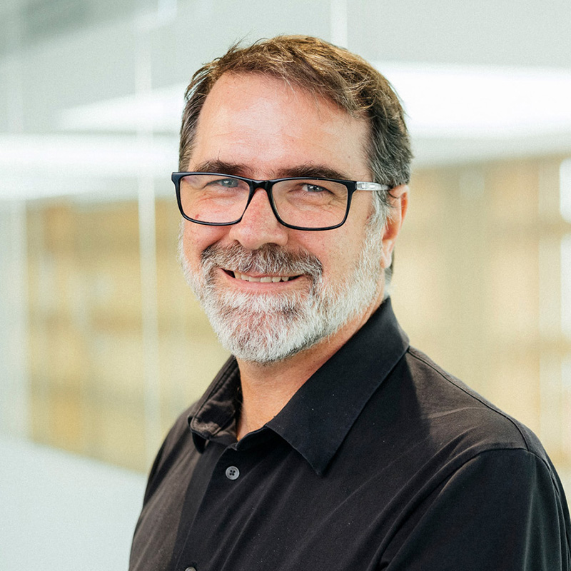 Picture of a white man with brown hair, a brown-white beard and glasses, with a black polo-shirt, against a white and beige background